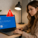 Notificaciones de Windows - Persona usando portátil con notificaciones de Windows: pantalla azul de error (BSOD) y icono de alerta rojo, fallo del sistema que requiere reiniciar el PC.