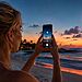 Fotos lunares con iPhone - Mujer fotografiando la luna llena con un iPhone en una playa al atardecer, aplicando trucos para captar sus detalles.